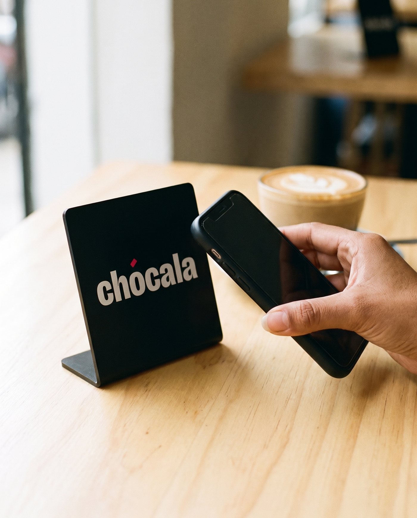 Mano acercando un iPhone al soporte de Chócala sobre la mesa de un café